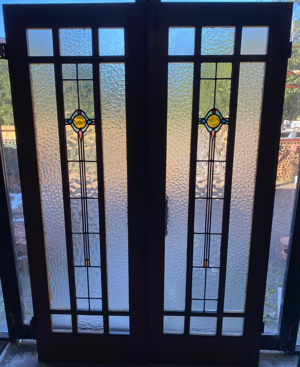 A pair of original salvaged 1920s timber French doors with three vertically stacked textured privacy glass window panels featuring subtle green and amber leadlight geometric motifs, shown in the Chippendale Restorations salvage workshop.