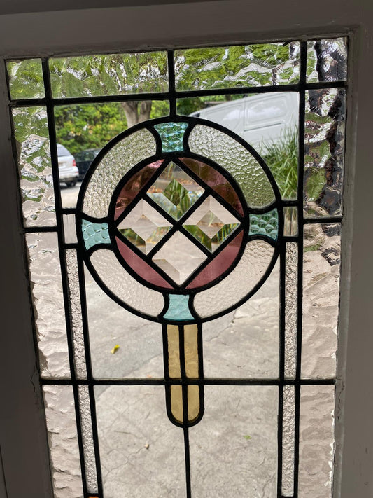 Close-up of the leadlight glass panel on one single casement window. The clear, heavily textured glass (likely "hammered" or "water glass") surrounds a central circular design composed of stained glass pieces in pastel pink, aqua, and pale yellow, surrounding a central clear glass bevel cluster. The black lead cames are visible.