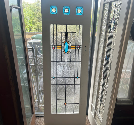 An original salvaged Federation style timber door painted white, featuring an upper window panel with green and amber bordered geometric leadlight privacy glass on display at Chippendale Restorations.