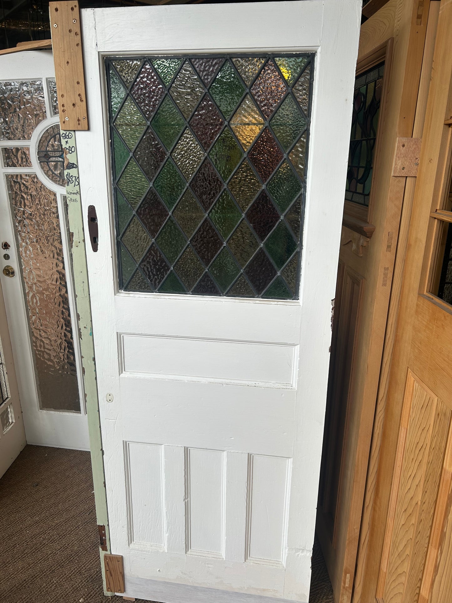An original salvaged timber Federation door painted white, featuring a large upper glass panel with green, purple, and amber diamond pattern leadlight stained glass inside the Chippendale Restorations warehouse.