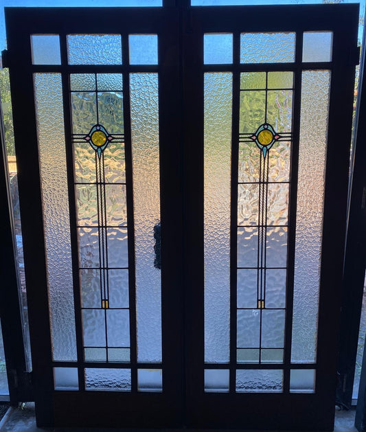 A pair of original salvaged 1920s timber French doors with three vertically stacked textured privacy glass window panels featuring subtle green and amber leadlight geometric motifs, shown in the Chippendale Restorations salvage workshop.