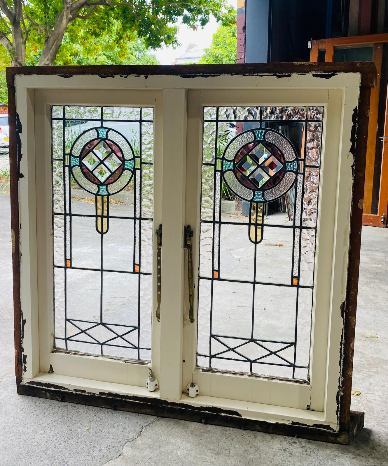 Antique double casement window set in a distressed white wooden frame with dark, peeling outer trim. The two matching leadlight panels feature a central circular stained glass design in pastel pink, aqua, and amber, surrounding a clear beveled diamond cluster. The rest of the panel uses clear, heavily textured glass with a lower geometric accent. Brass handles are visible in the center.