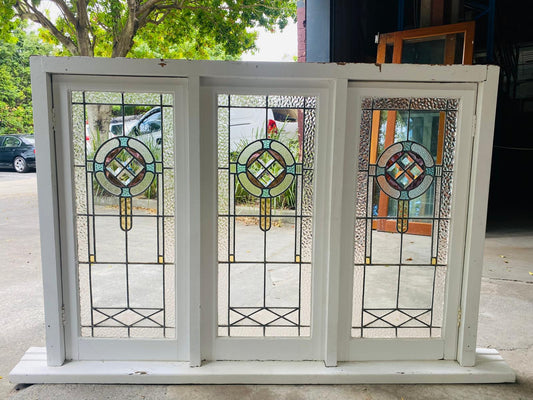 Large triple casement window unit in an off-white wooden frame with distressed dark trim. Each of the three leadlight panels features a matching central circular Art Deco/Art Nouveau motif in stained glass with pink, aqua, and amber tones, and a lower geometric accent panel. The clear glass is textured. The windows are displayed outdoors.