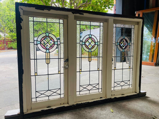 Full view of the triple casement leadlight window unit from the exterior. The white wooden frame surrounds three vertical panels. Each panel contains a decorative stained glass element with a circular design and lower cross-hatching motif, set against clear, heavily textured glass. The overall unit sits on a concrete floor with a white sill piece.