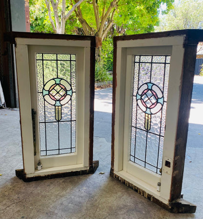 Two antique white wooden single casement windows with original, textured leadlight glass panels. Each panel features a central, circular Art Nouveau or Art Deco-style design in pale pink, blue, and amber stained glass, set against clear textured glass. The windows are propped open in a weathered wooden frame outdoors.
