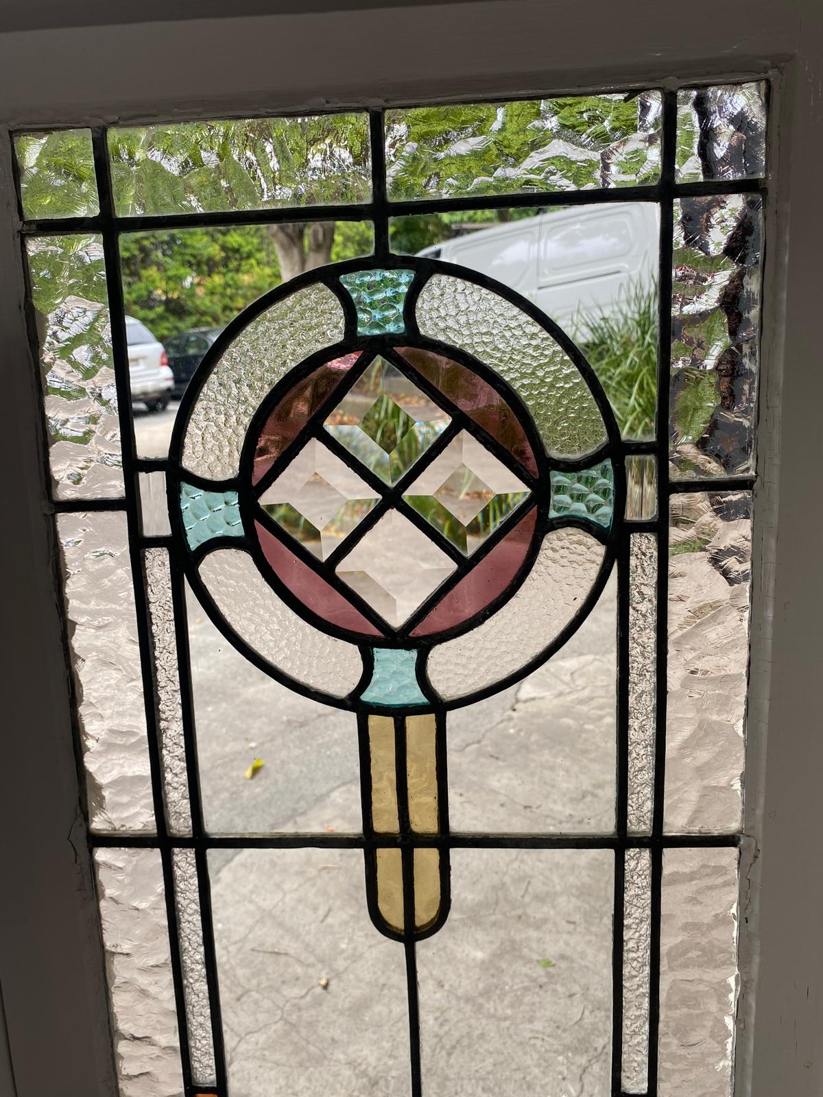 Close-up of the leadlight glass panel on one single casement window. The clear, heavily textured glass (likely "hammered" or "water glass") surrounds a central circular design composed of stained glass pieces in pastel pink, aqua, and pale yellow, surrounding a central clear glass bevel cluster. The black lead cames are visible.
