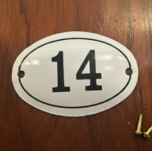 An oval white number plate with the black printed number 14, shown on a brown wooden surface with two mounting screws nearby.