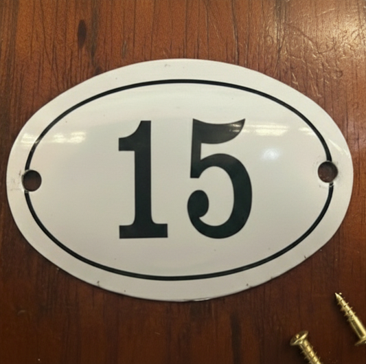 An oval white metal house number plaque with the number 15 in black, displayed on a brown wooden surface with two mounting screws nearby.