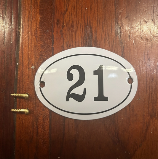 An oval white house number plaque with black numerals '21' and a black border, shown mounted on a brown wooden door with two gold screws beside it.