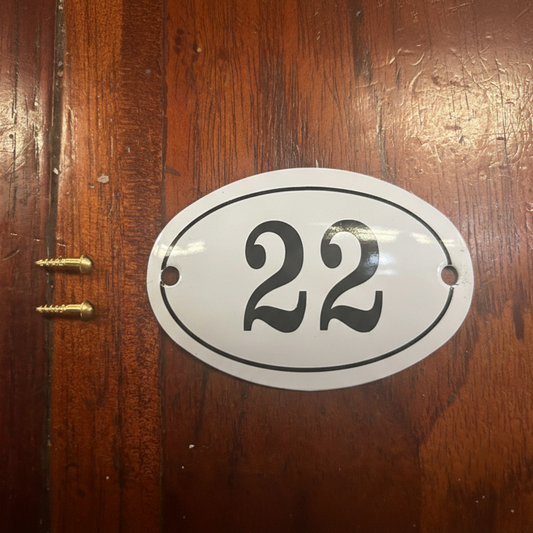 An oval white house number plate with black numerals '22' and a black border, shown mounted on a dark brown wooden door next to two gold-colored screws.