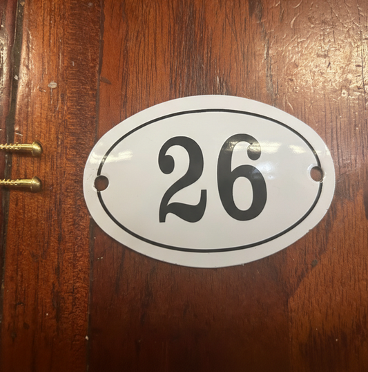An oval white house number sign with black border and the number 26, shown mounted on a dark brown wooden door.