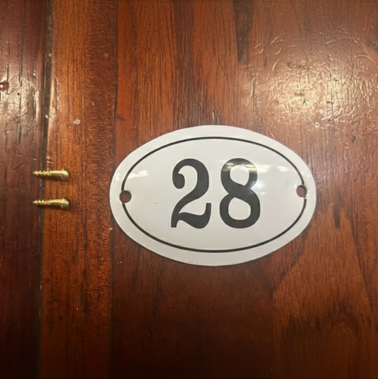 An oval white house number plaque with black numerals '28' and a thin black border, mounted on a dark brown wooden door.
