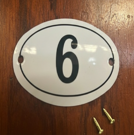 An oval white house number plaque with the number 6 in black, shown on a wooden surface with two mounting screws.