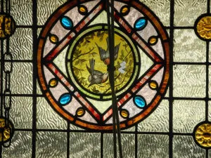 Close-up of an antique hand-painted glass roundel showing birds on branches inside a Victorian leadlight transom window frame.